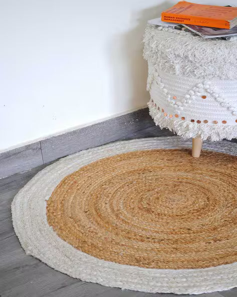 Round jute rug on a wooden floor with a white wall and textured white stool in the background.