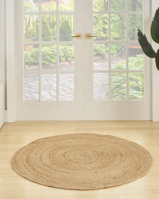 Round braided rug on a wooden floor in front of a glass door with a garden view.