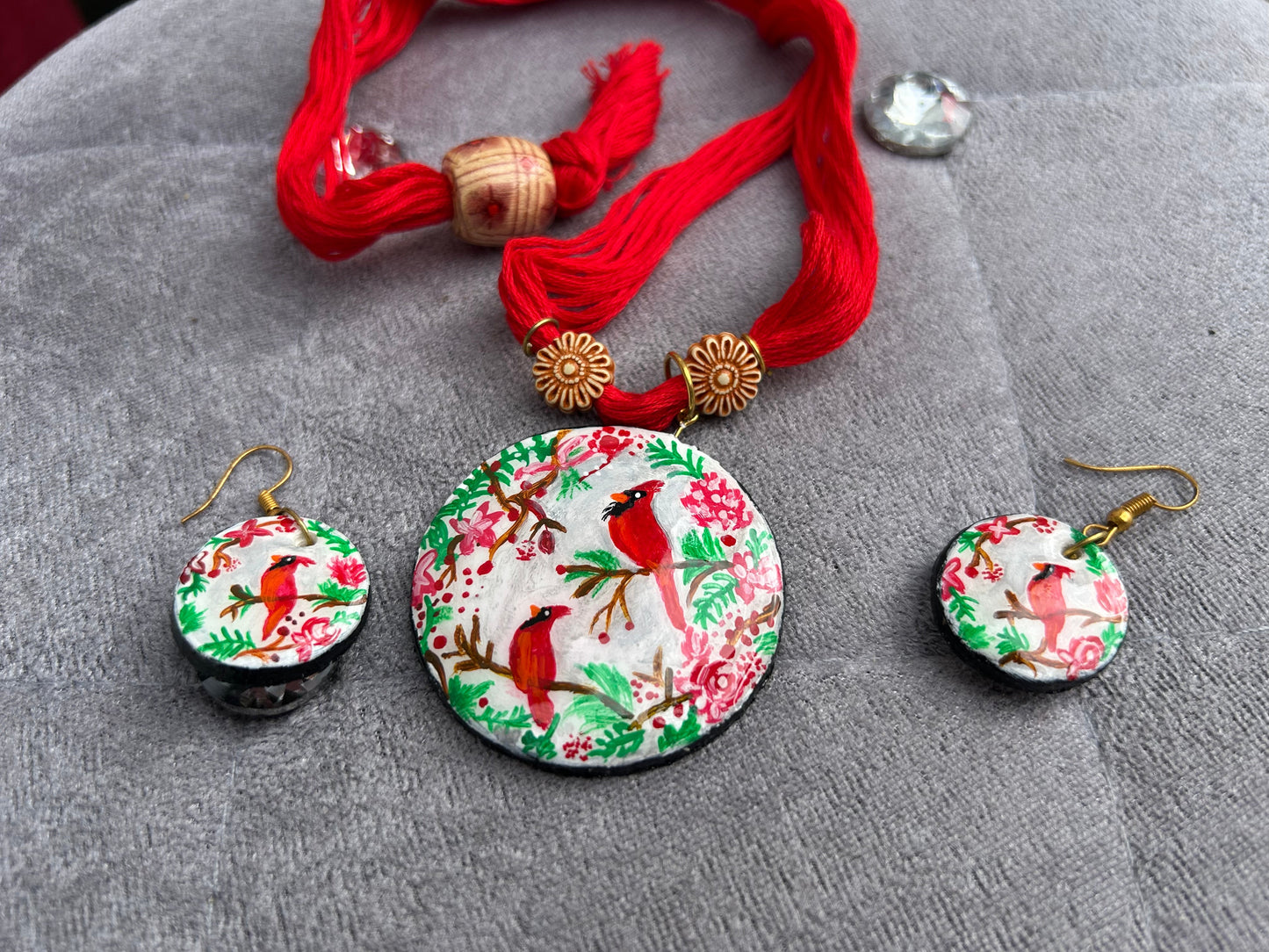 Jewelry set with pendant and earrings featuring red birds and floral patterns on a gray surface.