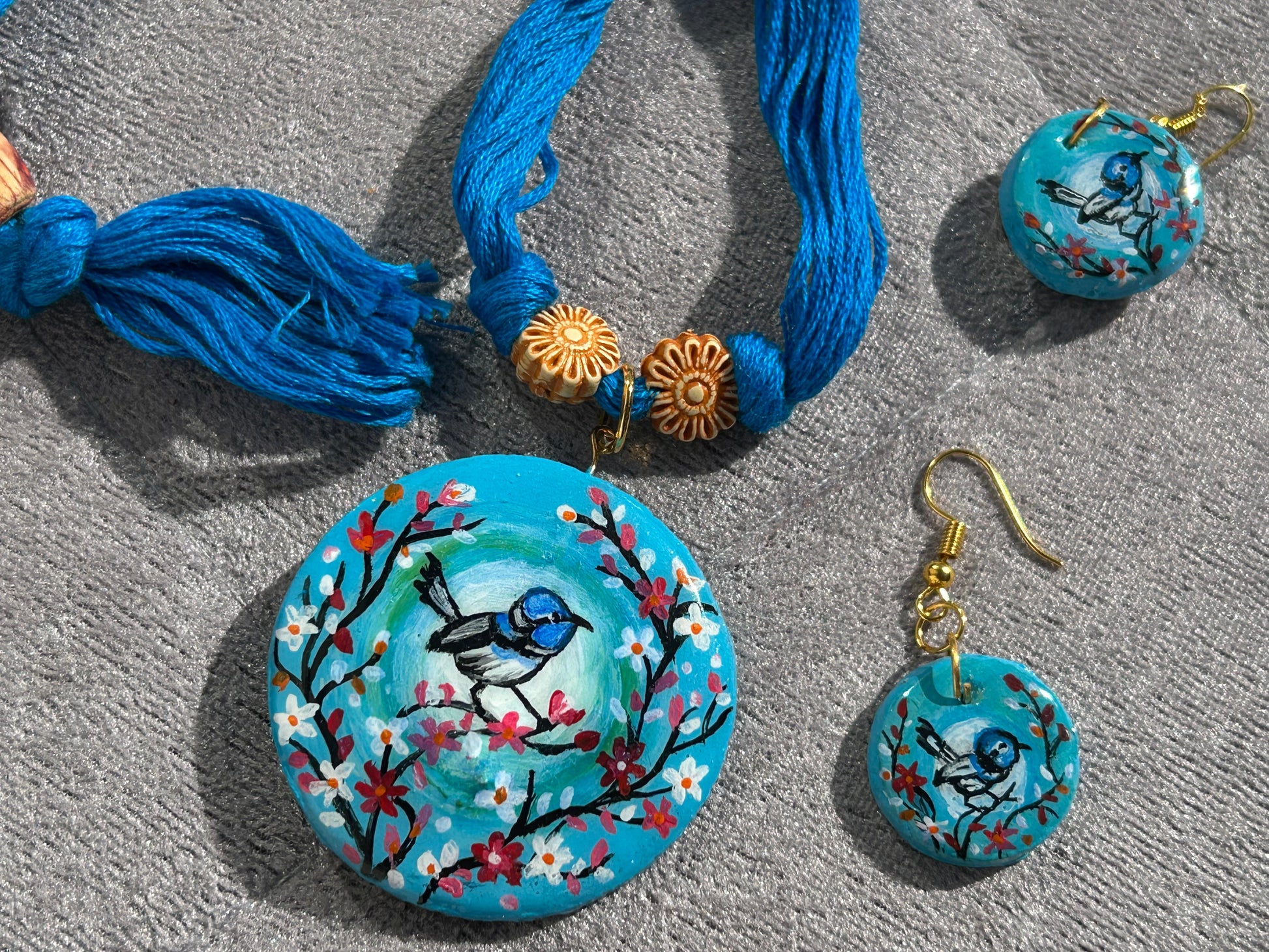 Jewelry set with blue necklaces and earrings featuring bird and floral designs on a gray surface.