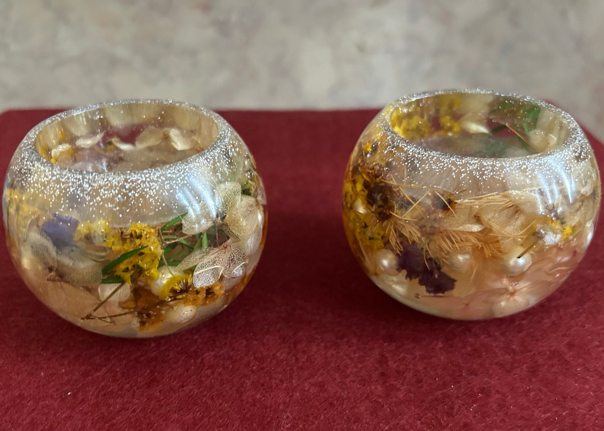 Two decorative glass containers with embedded flowers and pearls on a red surface.