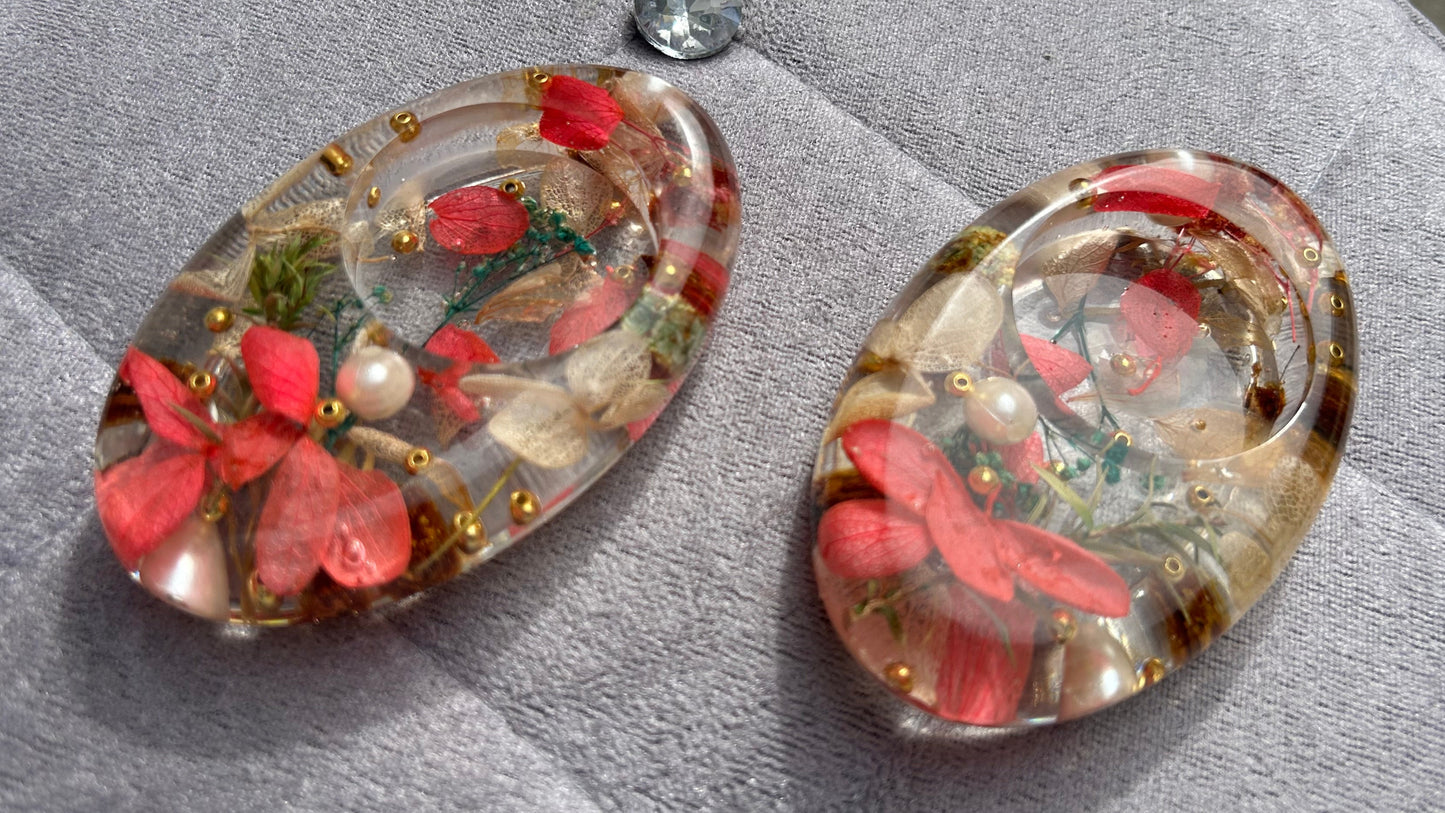Oval-shaped earrings with floral and pearl details on a textured surface.